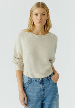 Licht beige pullovertrui met een geribbelde halslijn en mouwuiteinden, met een relaxte pasvorm en zachte textuur, gecombineerd met hooggeplaatste blauwe jeans.