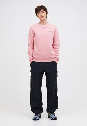 Person står med hænderne i lommerne, iført en lyserød sweatshirt, sorte cargobukser og grå træningssko mod en ensfarvet baggrund.