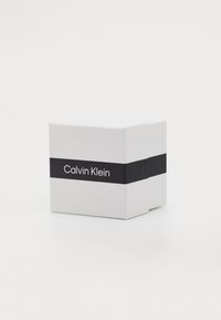 Boîte blanche carrée avec une bande noire affichant "Calvin Klein" en texte blanc. Fini mat lisse, design minimaliste.