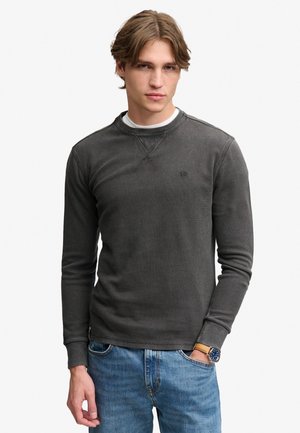 Ung mand med lyst brunt hår iført mørkegrå langærmet skjorte over hvid undertøj og blå jeans, med et ur på venstre håndled.