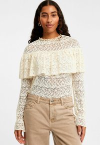 Lacebluse i cremefarve med flæseoverlap, lange ærmer og høj hals, parret med lysebrune denimbukser. Teksturopblomstermønster.