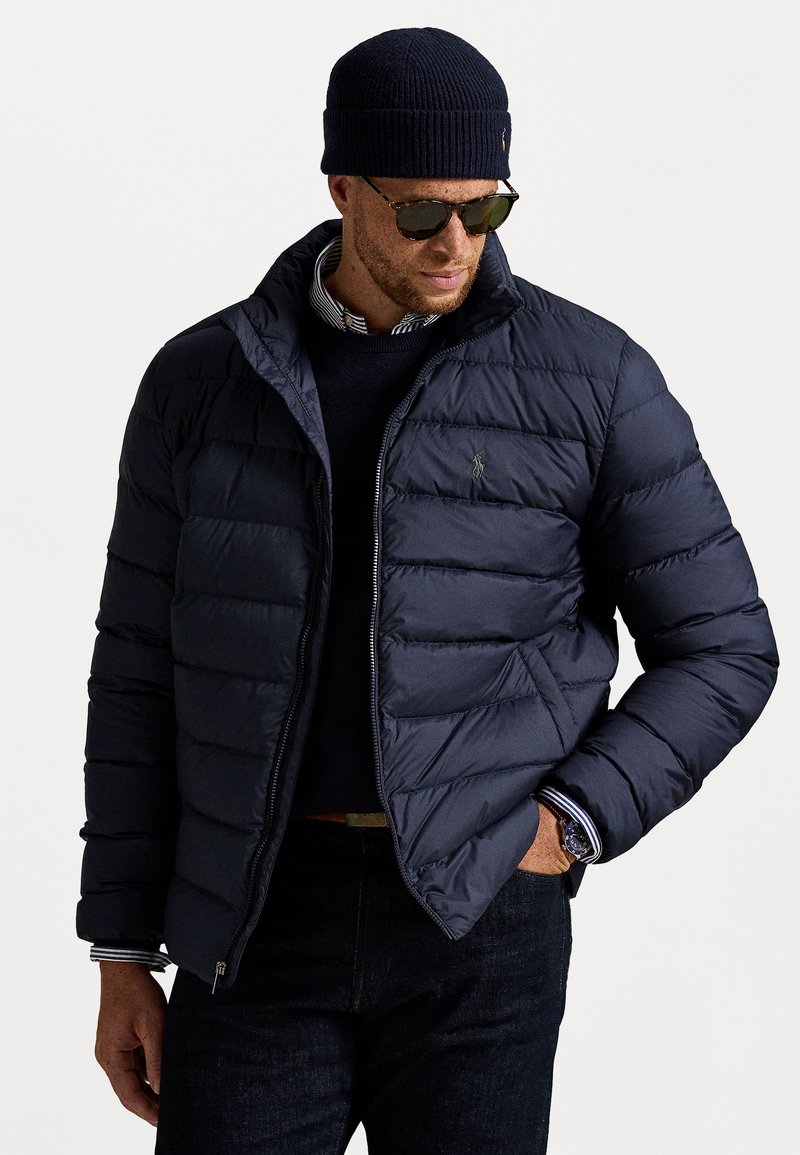 Mand iført en navy pufferjakke, stribet skjorte, sort sweater, mørke jeans, strikket hat, solbriller og ur, poserer med den ene hånd i lommen.