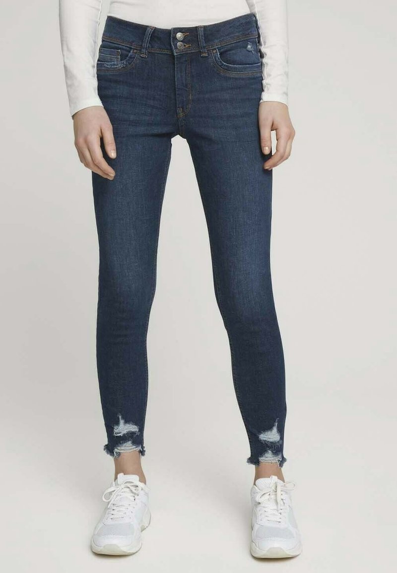 Mörkblå skinny jeans med fransat slut, fem fickor, knapp-stängning och lätt blekning; kombinerat med vita träningsskor.