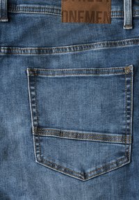 Blå denimjeans tyg med en ficka upptill till vänster, orange sömmar och ett brunt lädermärke med präglad text som är fäst i midjan.