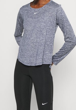 Person, die ein langärmliges, meliertes graues Sportoberteil und schwarze, enganliegende Leggings trägt, beide mit weißen Nike-Swoosh-Logos.