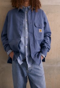 Blauwe button-up shirt gelaagd over een lichterblauwe denim shirt, beide met klassieke kragen; losse denim jeans; voorzien van een logo patch op de bovenste zak.