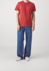 Rød bomulds T-shirt med rund hals og et lille logo på brystet, sammen med brede blå denim jeans og hvide sneakers.