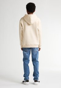 Felpa beige con cappuccio, vestibilità rilassata, texture morbida. Jeans in denim blu, gamba dritta, lavaggio sbiadito. Sneakers bianco e nere con accenti gialli.