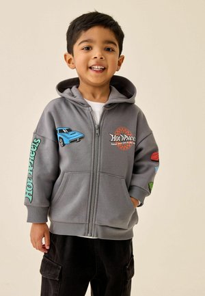 Young boy smiling, wearing grey Hot Wheels zip-up hoodie with car graphics and black cargo pants, standing against plain background.