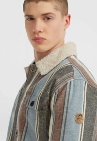 Jeune homme portant une veste rayée multicolore avec un col en shearling blanc, tourné légèrement sur le côté avec une expression neutre.