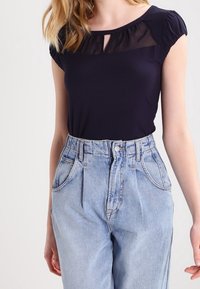 Mujer con una blusa negra de manga corta y yugo transparente, y unos vaqueros de mezclilla azul claro de cintura alta, de pie frente a un fondo liso.