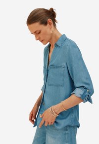 Camicia di jeans azzurro chiaro con maniche lunghe arrotolate, dotata di due tasche sul petto e colletto. Tessuto liscio e vestibilità rilassata, abbinata a jeans in denim.