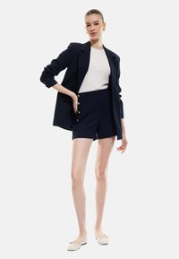 Blazer navy con maniche arrotolate, top bianco a coste e pantaloni navy con dettagli a bottoni, abbinati a scarpe ballerina chiare. Design semplice e strutturato.