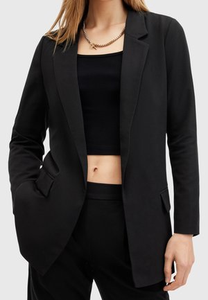 Frau trägt schwarzen maßgeschneiderten Blazer über einem kurzen gerippten Top und passenden schwarzen Hosen, accessoriert mit einer massiven goldenen Kettencollier.