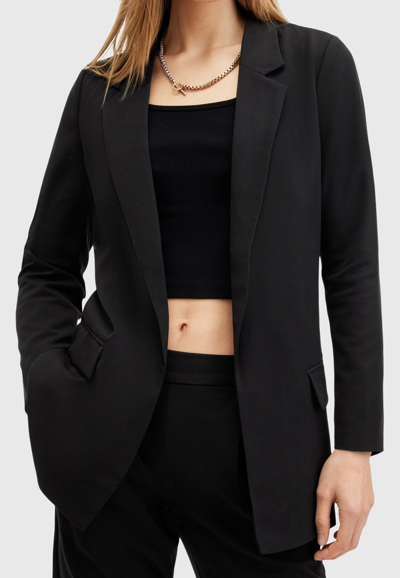 Femme portant un blazer noir tailleur sur un top court côtelé et un pantalon noir assorti, accessoirisée d'un collier chaîne en or massif.