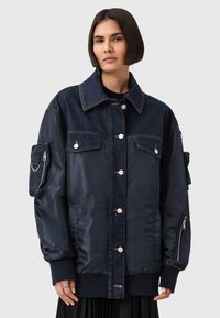 Donna che indossa un giubbotto sovradimensionato blu scuro che combina denim e nylon, con chiusura a bottoni, tasche con zip sulle maniche e polsini ripiegati.