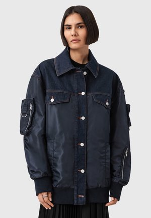 Donna che indossa un giubbotto sovradimensionato blu scuro che combina denim e nylon, con chiusura a bottoni, tasche con zip sulle maniche e polsini ripiegati.