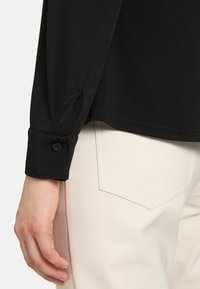 Black long-sleeve blouse with a smooth texture and buttoned cuffs, paired with light-colored pants featuring a back pocket detail.