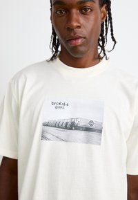 Light cream cotton t-shirt featuring a black-and-white graphic photo print with the word "DICKIES" and truck imagery. Short sleeves, round neckline.