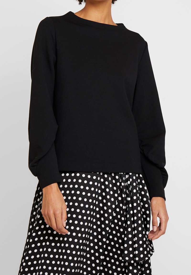 Femme portant un haut noir à manches longues associé à une jupe noire à pois blancs avec un détail de volants asymétrique.