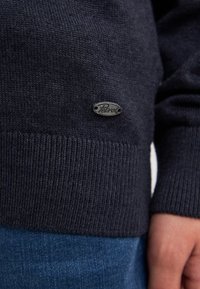 Marineblauer Strickpullover mit geripptem Saum und strukturiertem Finish. Verfügt über ein kleines silbernes Logoetikett an der unteren Seite, kombiniert mit Jeans.