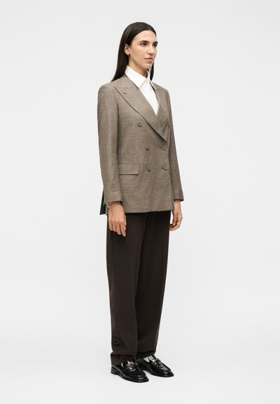 Γυναίκα με μακριά σκούρα μαλλιά, ντυμένη με καφέ διπλόμπελο σακάκι, λευκό πουκάμισο, φαρδιά σκούρα παντελόνια και μαύρα μοκασίνια, στέκεται πλάγια.