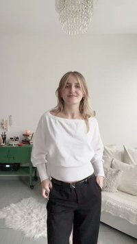 Witte, loszittende trui met geribde zoom; gecombineerd met zwarte, wijd uitlopende broek. Minimalistisch ontwerp, gladde textuur, zonder opvallende patronen.