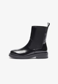Bottines Chelsea noires en matériau brillant avec des panneaux élastiques sur les côtés, bout rond et semelle noire épaisse. Onglet en boucle à l'arrière pour un enfilage facile.