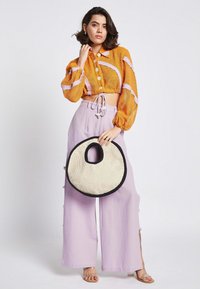 Camicetta arancione corta con righe lavanda, abbinata a pantaloni larghi lavanda. Tiene in mano una borsa rotonda intrecciata con bordi neri.
