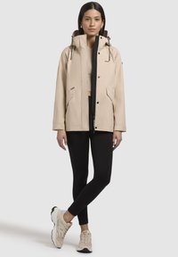 Beige waterdichte jas met een hoge kraag, voorzakken en zwarte accenten, gecombineerd met zwarte leggings en lichte sneakers.