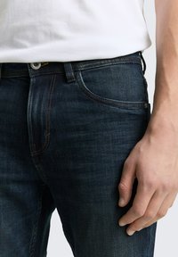 Ciemnoniebieskie dżinsy z denimu o dopasowanym kroju, wyposażone w klasyczny design z pięcioma kieszeniami, widocznymi przeszyciami oraz zapięciem na guzik w pasie.