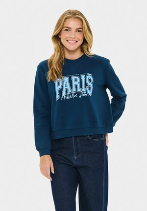 Femme souriante avec de longs cheveux blonds, portant un sweat bleu avec le texte "PARIS Athletic Dept." et un jean bleu foncé, se tenant devant un fond uni.