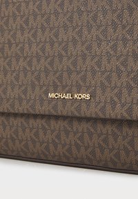 Brown patterned textile handbag featuring a gold "Michael Kors" logo. The design includes a geometric pattern and a structured shape.