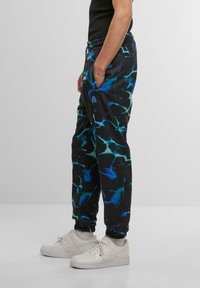 Sort træningsbukser med et blåt og grønt abstrakt mønster, elastisk talje og manchetter. Båret med hvide sneakers.