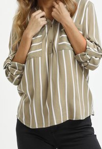 Personne portant une chemise à manches retroussées beige et blanche à rayures verticales, tenant le col de la chemise, associée à un pantalon noir.