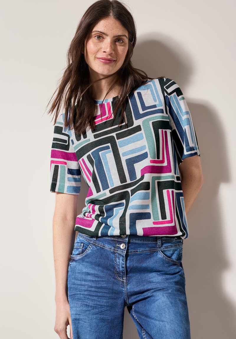 Haut multicolore à manches courtes présentant des motifs géométriques en bleu, rose, vert et noir ; fabriqué en tissu doux, associé à un jean bleu.