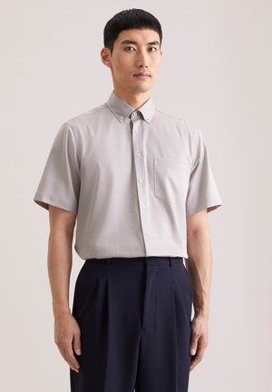 Uomo che indossa una camicia grigio chiaro a maniche corte con taschino sul petto e pantaloni scuri a pieghe, davanti a uno sfondo semplice.