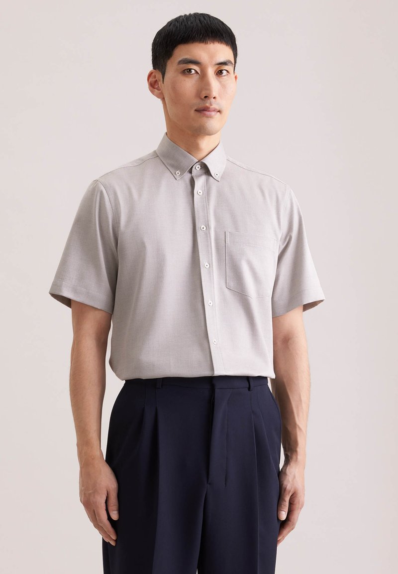 Homme portant une chemise à manches courtes gris clair avec poche poitrine et un pantalon plissé foncé, devant un fond uni.