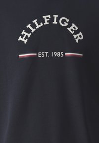 T-shirt di cotone blu navy con testo bianco "HILFIGER" ad arco sopra "EST. 1985" e strisce orizzontali rosse e bianche sotto. Texture liscia.