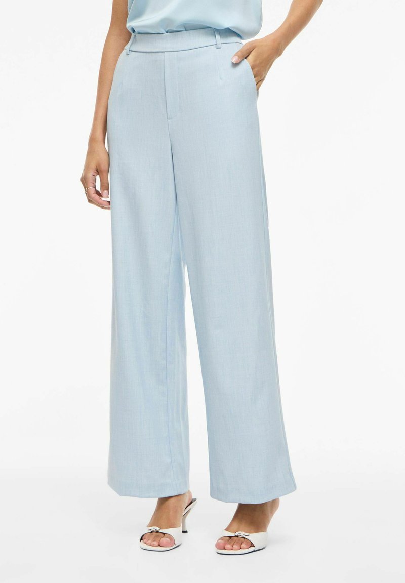 Žena oblečená v světle modrých širokých kalhotách a bílých sandálech s otevřenou špičkou stojí s jednou rukou v kapse na bílém pozadí.