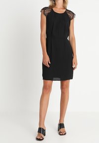 Černé šaty s krátkými krajkovými rukávy, kulatým výstřihem a vypasovaným pasem. Hladký materiál se sukní do délky kolen. Nosené s černými podpatky.