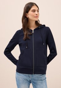 Haut zippé bleu marine en tissu doux, avec une poche kangourou, une capuche à cordon de serrage et des poignets côtelés. Design décontracté avec une texture lisse.