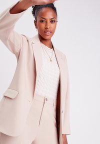 Beige tailored blazer with notch lapels, paired with a textured white top. Model raises hand, showcasing a simple pendant necklace.