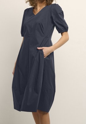 Femme portant une robe bleu marine longueur genou avec des manches courtes bouffantes et un décolleté en V, debout avec une main dans une poche latérale.