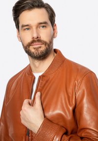 Giacca bomber in pelle marrone con colletto e polsini a coste. Chiusura frontale con zip, trama liscia, design aderente, mostrata mentre è tenuta da un modello.