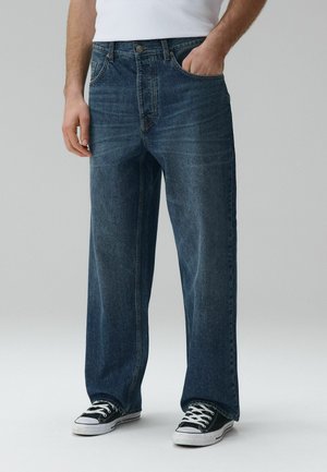 Mężczyzna w luźnych ciemnoniebieskich dżinsach i czarnych tenisówkach z białymi sznurowadłami, stojący z jedną ręką w kieszeni.