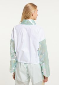 Iridescerende vindjakke med en hvid krop og skinnende lyseblå ærmer. Har en høj krave og elastiske manchetter. Letvægtigt stof.