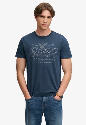 Młody mężczyzna o jasnobrązowych włosach, ubrany w niebieską koszulkę Superdry Co. i niebieskie dżinsy, z rękami w kieszeniach, na jednolitym jasnym tle.