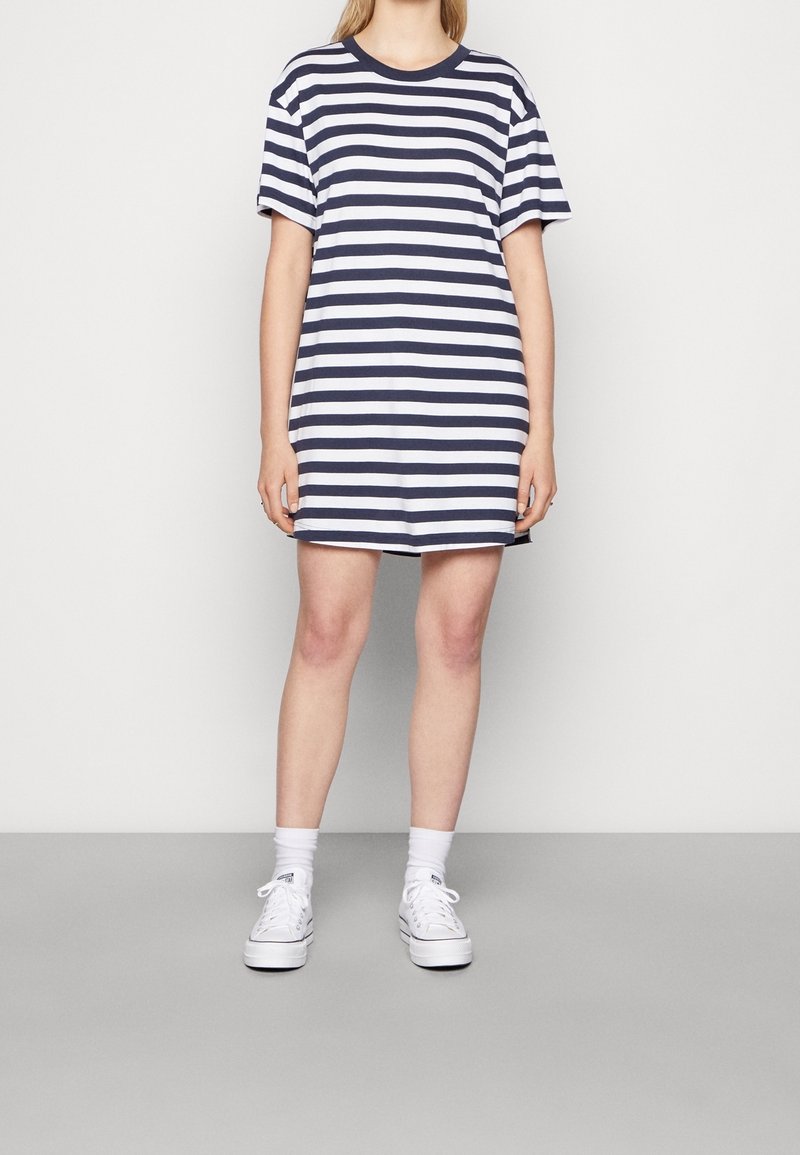 Raitapaita-mekko tummansinisenä ja valkoisena, valmistettu pehmeästä puuvillasta. Ominaisuuksiin kuuluu pyöreä kaula-aukko ja rento malli, joka ulottuu reiden puoliväliin. Valkoiset tennarit.