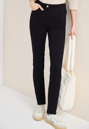 Personne portant un pantalon noir slim et des baskets blanches, tenant un sac fourre-tout matelassé blanc d'une main, l'autre main dans la poche.